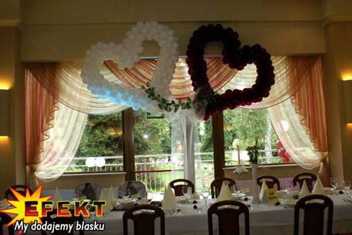 Dekoracja sali balonami w kształcie serc: białe z podświetleniem i bordowe, zawieszone nad stołem z zastawą, w tle okno i zasłony. Na pierwszym planie logo firmy.