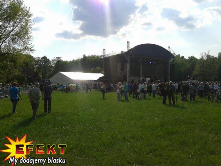 Plenerowa impreza masowa z dużą sceną i białym namiotem w tle, otoczona zielenią i zgromadzonym tłumem ludzi w słoneczny dzień. Logo firmy w rogu zdjęcia.