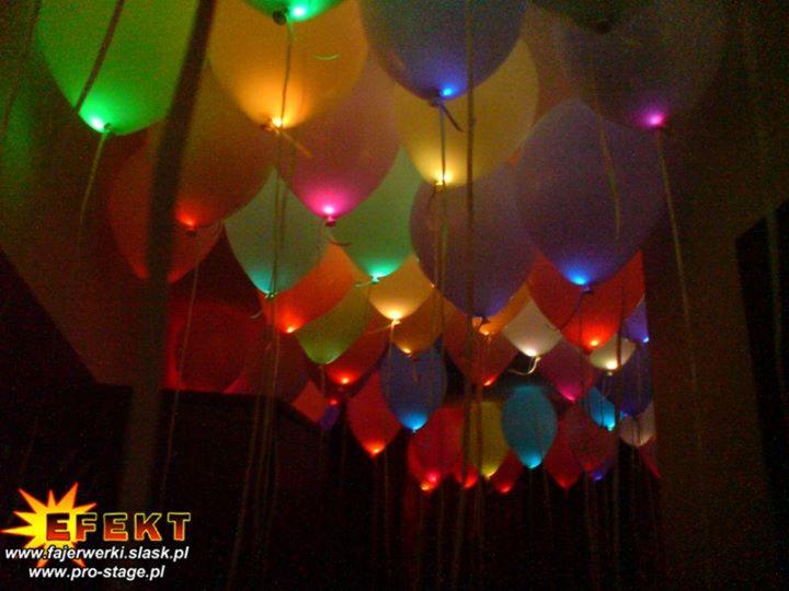 Kolorowe balony z podświetleniem LED, tworzące dekorację sufitową. Balony zawieszone na cienkich sznurkach, w tle ciemne otoczenie. Widoczne logo firmy w lewym dolnym rogu.