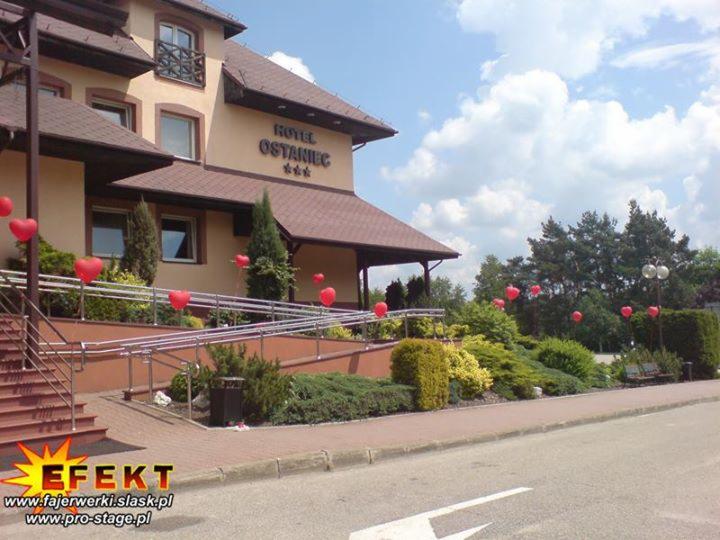 Hotel Ostaniec udekorowany czerwonymi balonami w kształcie serc, prawdopodobnie z okazji wydarzenia. Widoczny budynek, zieleń i logo firmy w rogu.