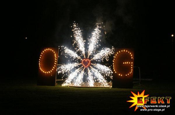 Pokaz pirotechniczny z efektem serca, flankowany literami 'O' i 'G' podświetlonymi punktowymi światłami, na tle nocnego nieba. Efekt specjalny na imprezie.