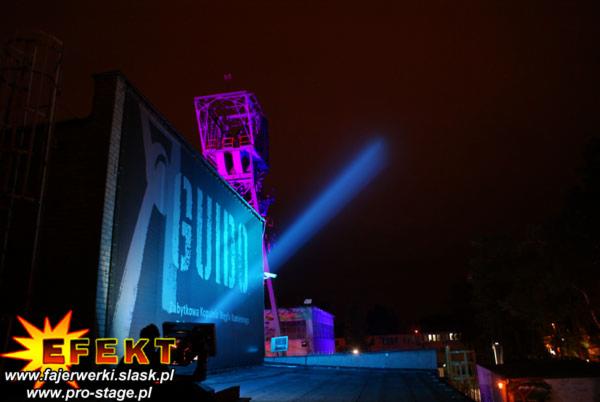 Nocna iluminacja budynku z napisem i wieżą, oświetlona niebieskim światłem. Zastosowano efekty świetlne i iluminację architektoniczną.