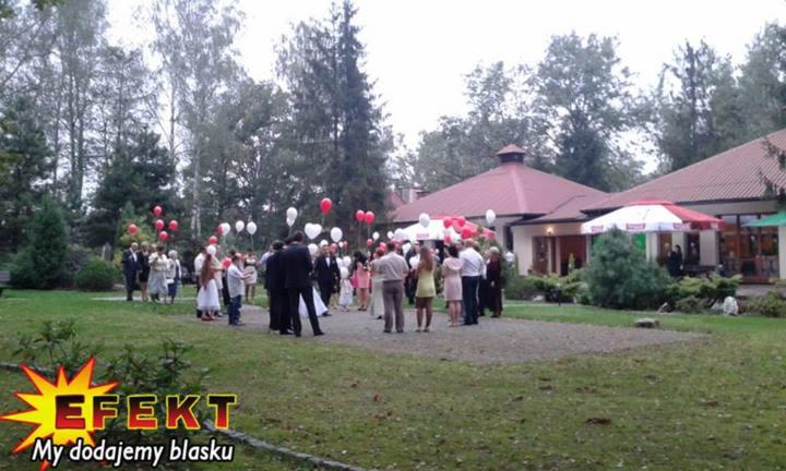 Grupa ludzi z balonami przed budynkiem z czerwonym dachem. Na pierwszym planie trawnik i logo firmy z napisem 'My dodajemy blasku'.