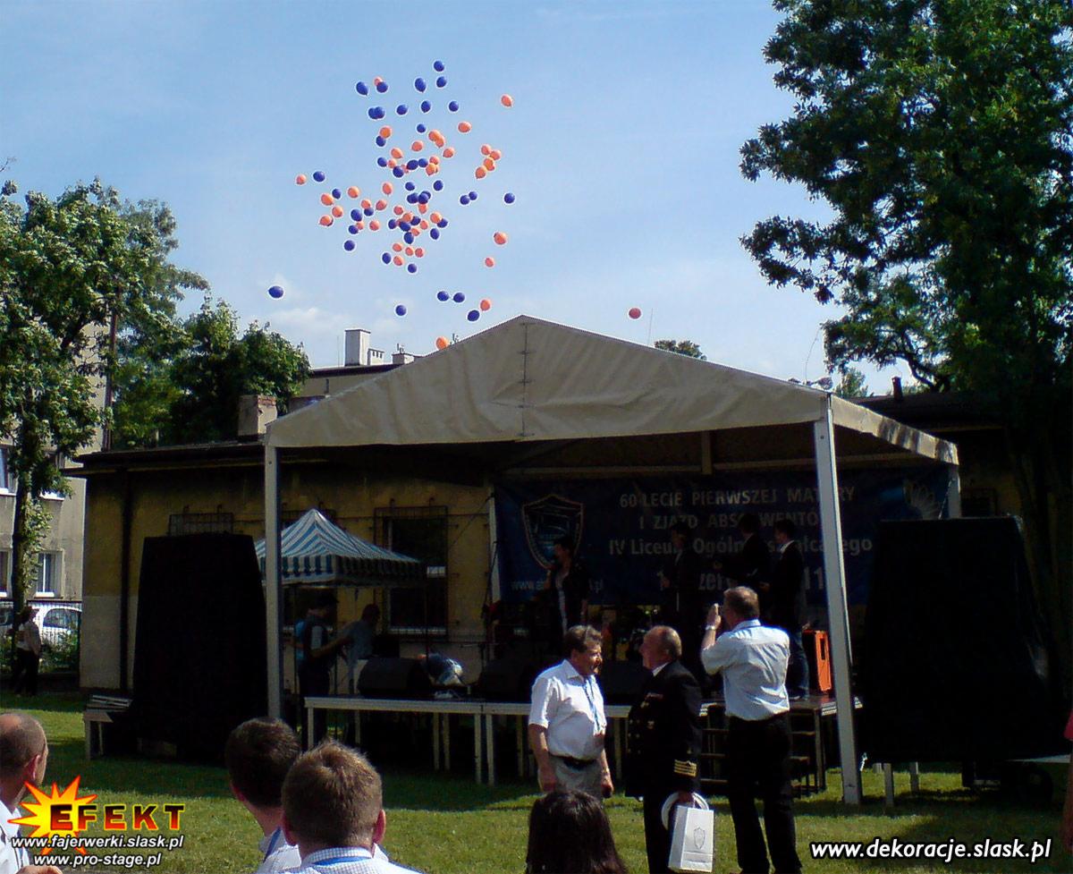Uroczystość na zewnątrz pod namiotem z balonami, z widoczną sceną i publicznością. Obchody 60-lecia szkoły, z elementami nagłośnienia.