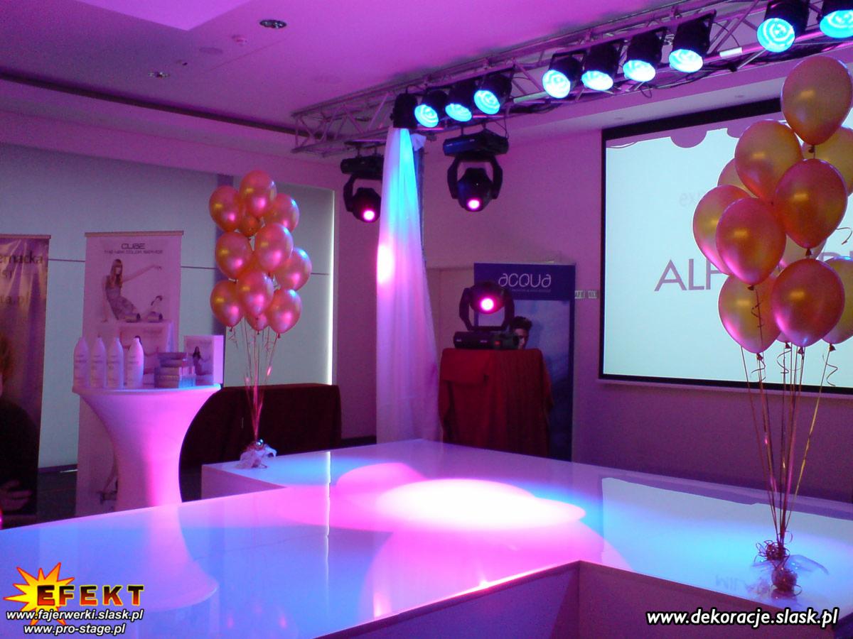 Scena oświetlona fioletowymi reflektorami z balonami po bokach. W tle ekran projekcyjny i stoisko z kosmetykami. Uwagę przykuwa błyszcząca powierzchnia sceny.