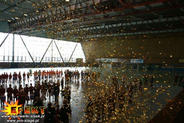 Konfetti opada na grupę ludzi w hali sportowej podczas wydarzenia. Widoczny baner firmy z efektami specjalnymi.