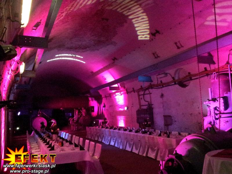 Długa sala bankietowa z białymi stołami i krzesłami, oświetlona różowym światłem, z projektorem wyświetlającym napisy na sklepieniu w industrialnym wnętrzu.