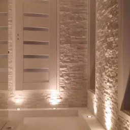 Nowoczesna łazienka z oświetleniem punktowym, ścianami wyłożonymi dekoracyjną płytką imitującą kamień i białymi drzwiami z przeszkleniami.