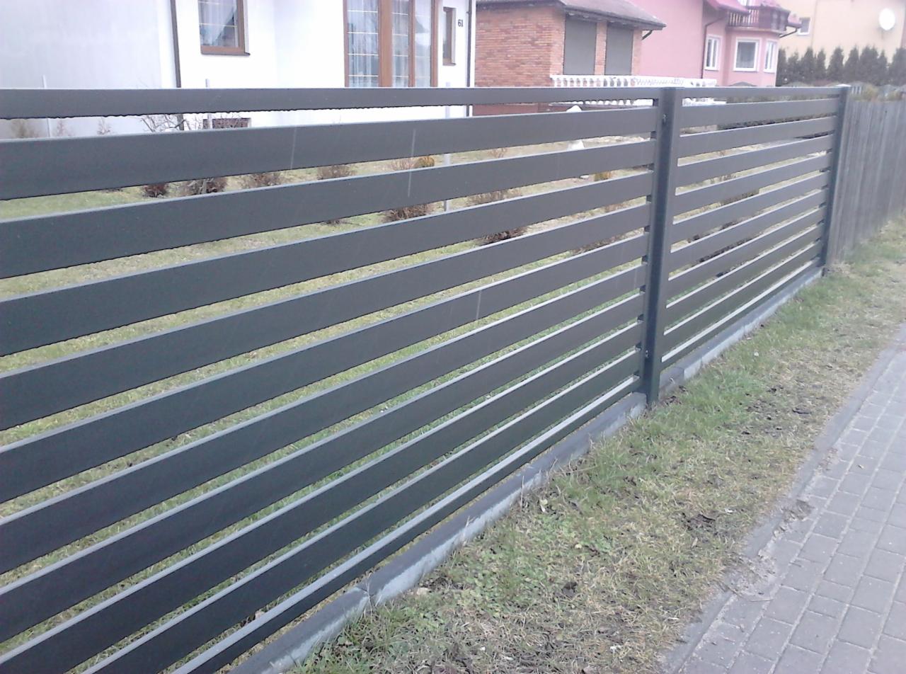 Nowoczesne przęsła ogrodzeniowe z poziomych, szarych profili metalowych, zamontowane na betonowej podmurówce, widok z zewnątrz posesji.