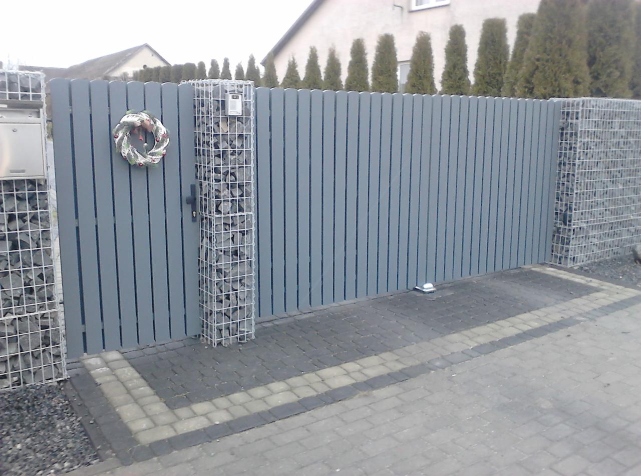 Szara brama z pionowych desek, wsparta na słupkach z siatki gabionowej wypełnionej kamieniami; na bramie zawieszony wieniec świąteczny, a obok skrzynka pocztowa.