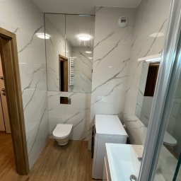 Adam Antonowski Remonty Mieszkań - Nowoczesna łazienka z marmurowymi płytkami, drewnopodobną podłogą, toaletą, pralką i umywalką z szafką, widoczna przez częściowo otwarte drzwi z drewnianą framugą.