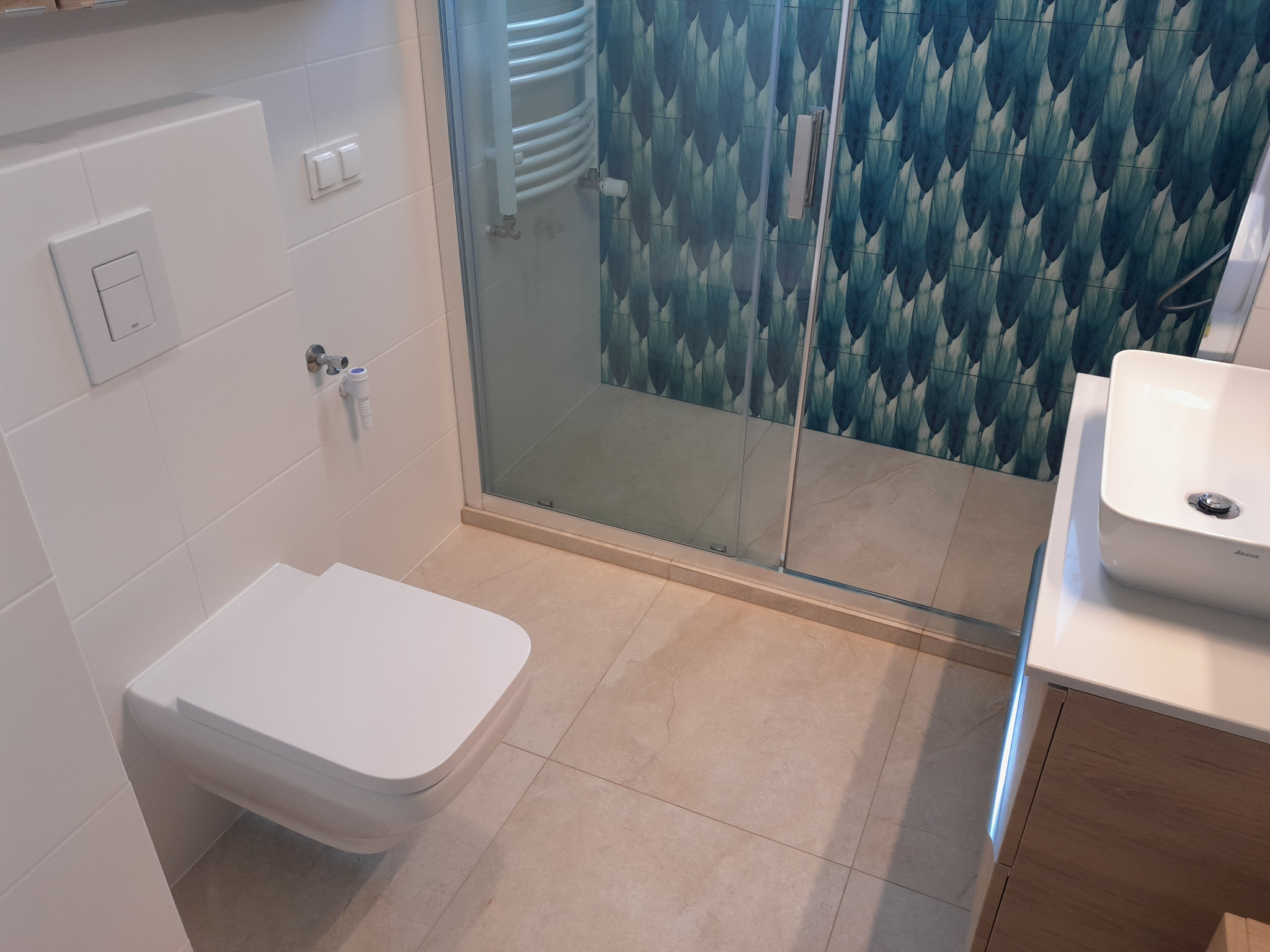 Nowoczesna łazienka z podwieszaną toaletą, kabiną prysznicową z zielonymi płytkami w liście, umywalką nablatową i beżowymi płytkami podłogowymi.