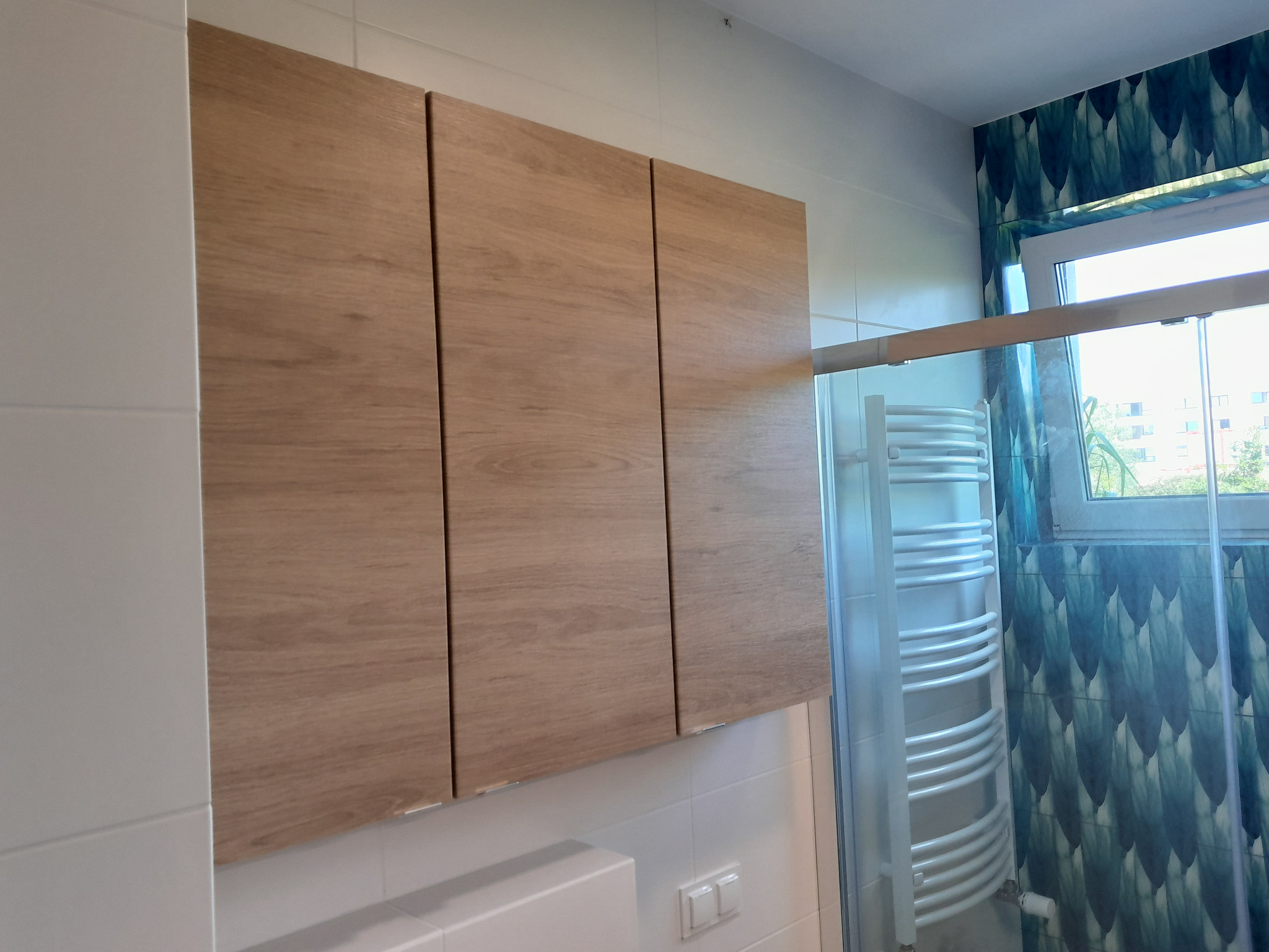 Nowoczesna łazienka z drewnianymi szafkami wiszącymi, białymi płytkami i grzejnikiem drabinkowym za szklaną kabiną prysznicową, tapeta w odcieniach niebieskiego z motywem liści przy oknie.