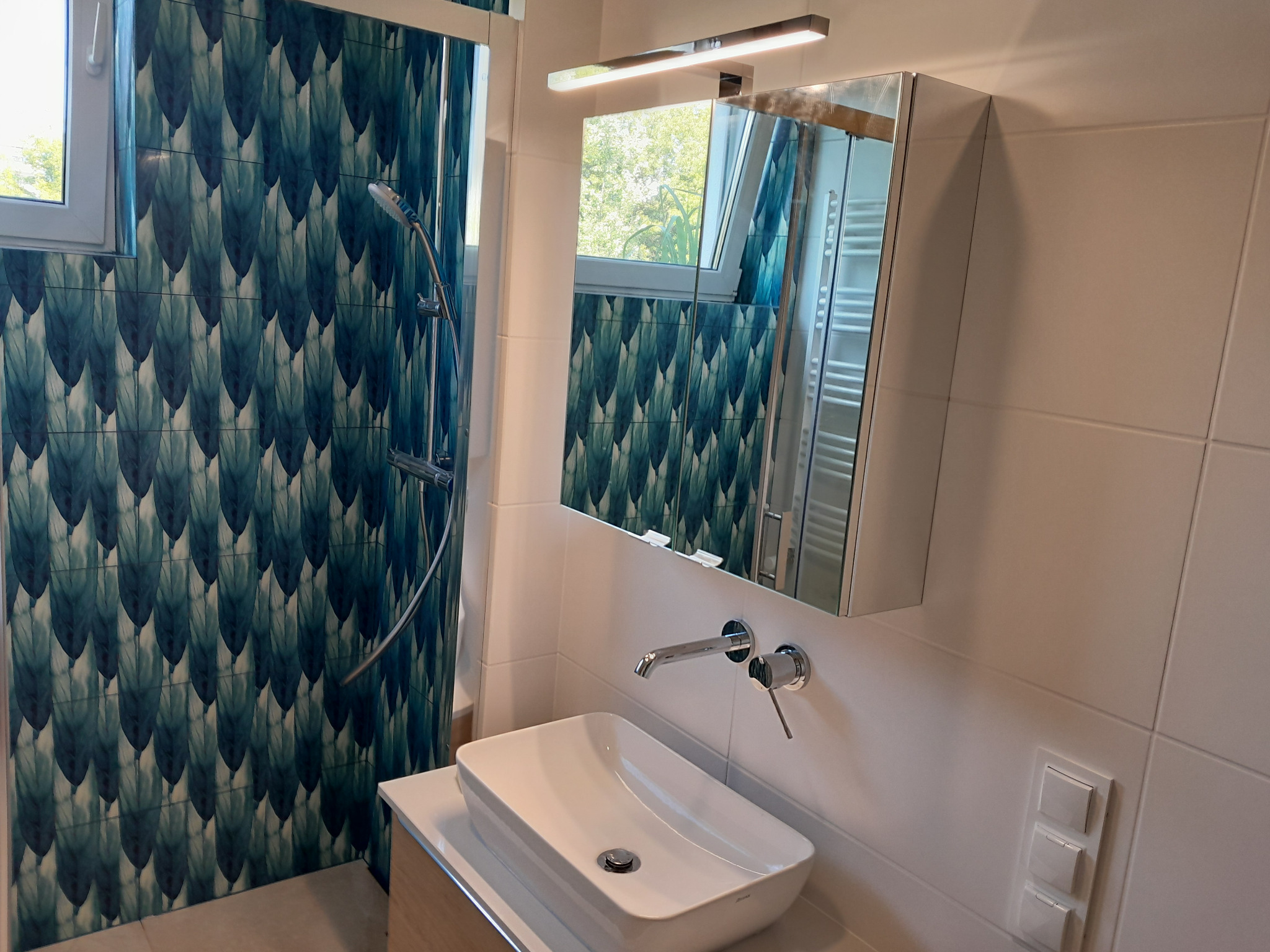 Nowoczesna łazienka z umywalką nablatową, baterią ścienną i lustrem z oświetleniem, akcentem są płytki w kabinie prysznicowej z wzorem w odcieniach zieleni i błękitu.