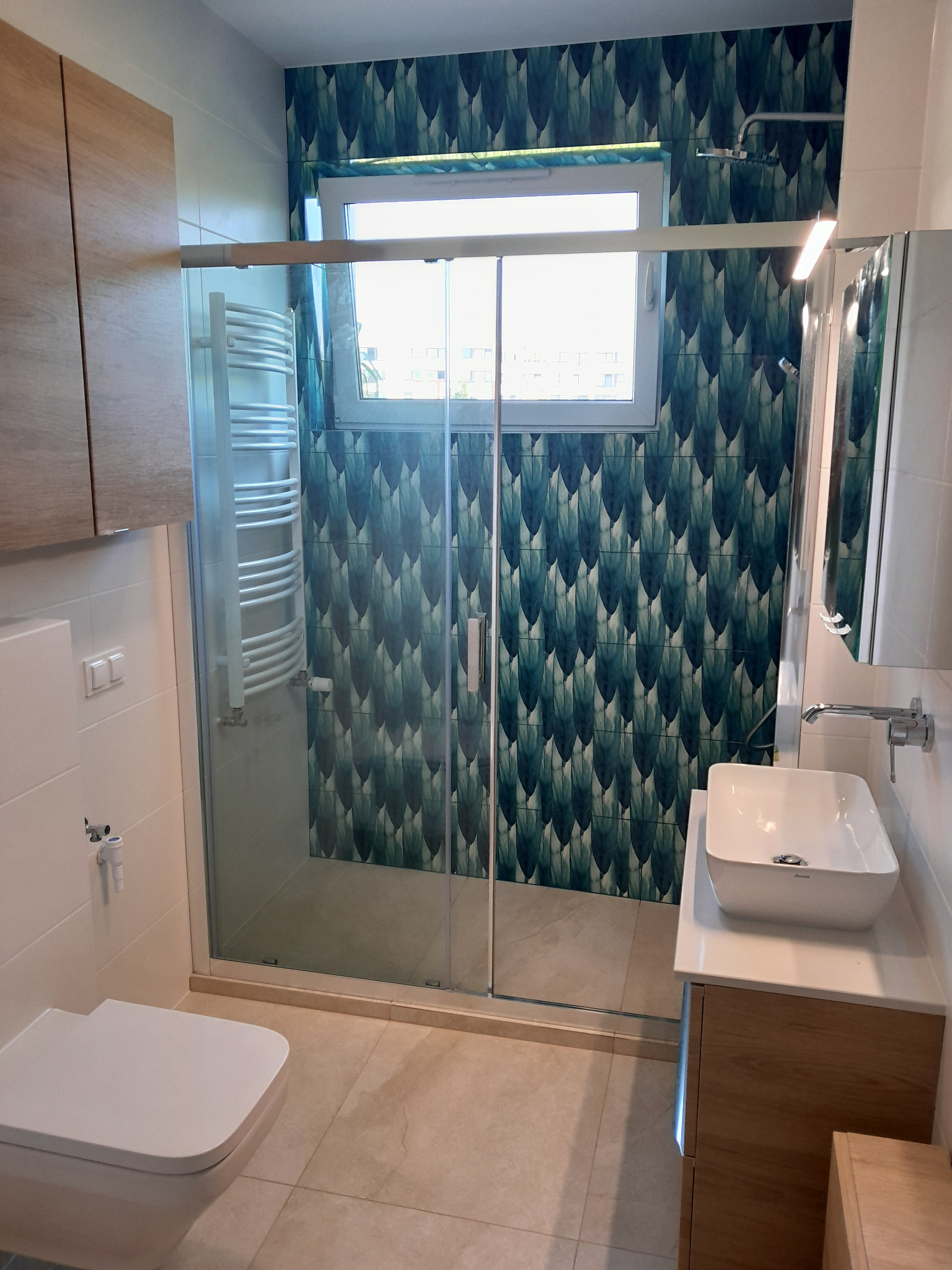 Nowoczesna łazienka z kabiną prysznicową typu walk-in, umywalką nablatową na szafce z drewnopodobnym frontem, toaletą i grzejnikiem drabinkowym; ściana za kabiną pokryta tapetą w motywy roślinne.