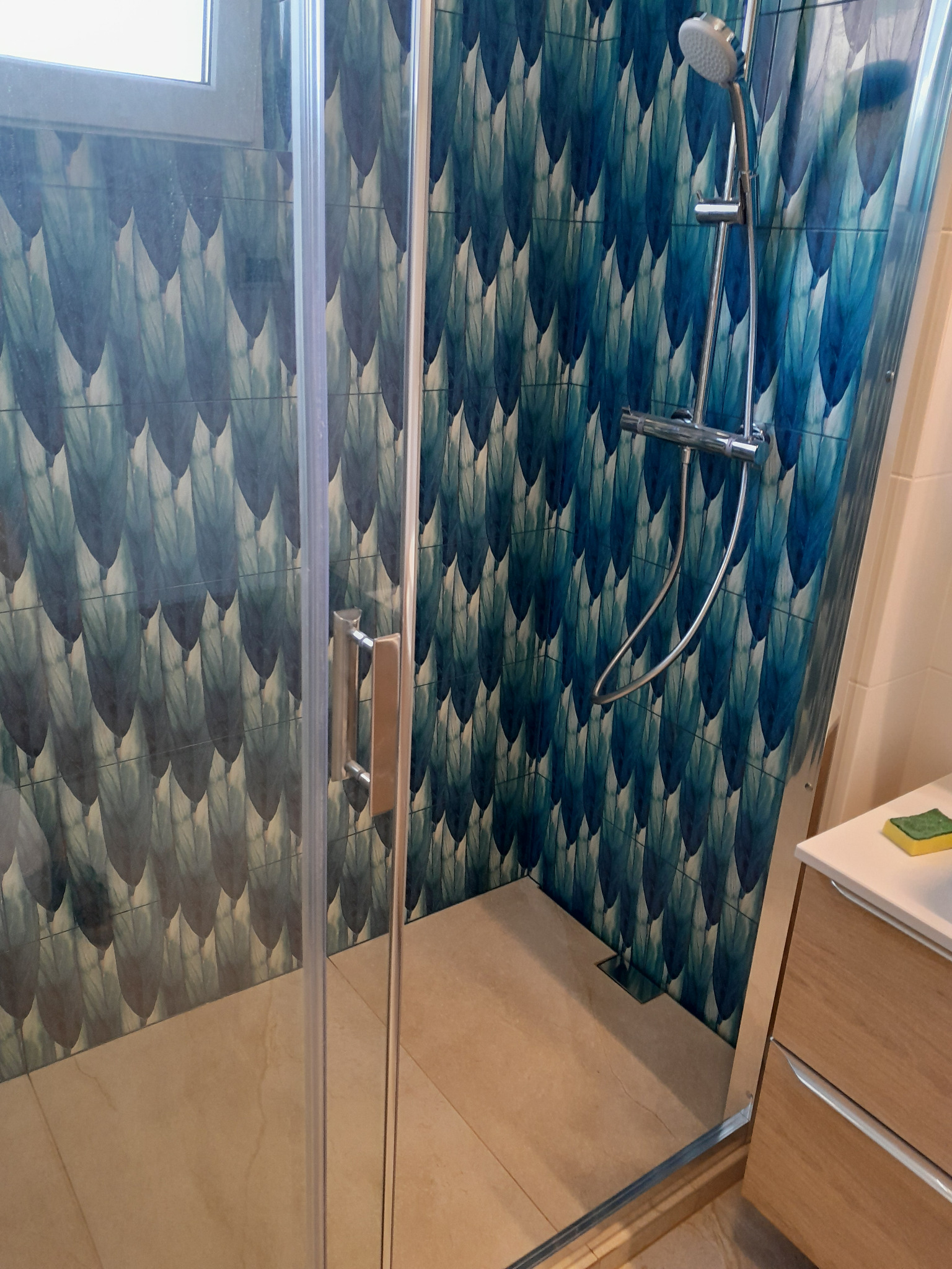 Kabina prysznicowa z niebieskimi płytkami w kształcie piór, chromowana armatura, beżowa podłoga i fragment szafki łazienkowej z blatem.