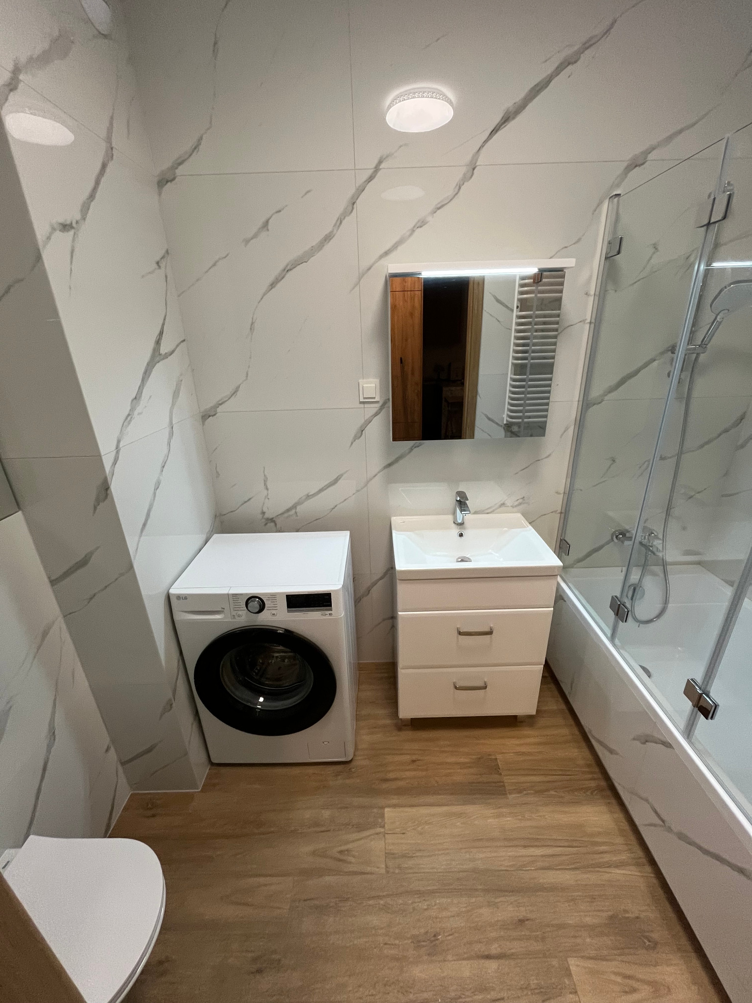 Nowoczesna łazienka z białymi płytkami imitującymi marmur, drewnopodobną podłogą, białą pralką LG, umywalką z szafką i kabiną prysznicową.