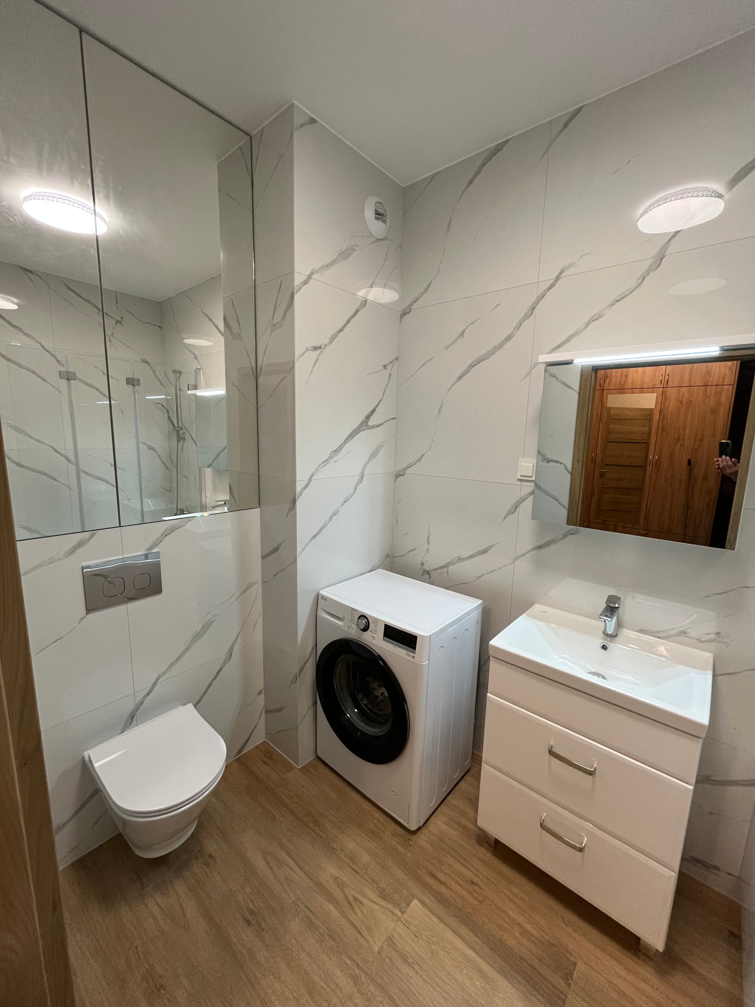 Nowoczesna łazienka z płytkami imitującymi marmur, wiszącą toaletą, pralką i szafką z umywalką, oświetlona ledowym plafonem i lustrem z podświetleniem.