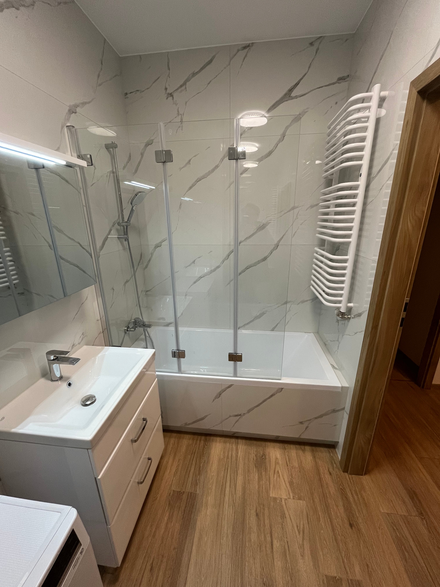 Nowoczesna łazienka z białą umywalką nablatową, kabiną prysznicową z transparentnego szkła, grzejnikiem drabinkowym i płytkami imitującymi marmur na ścianach.