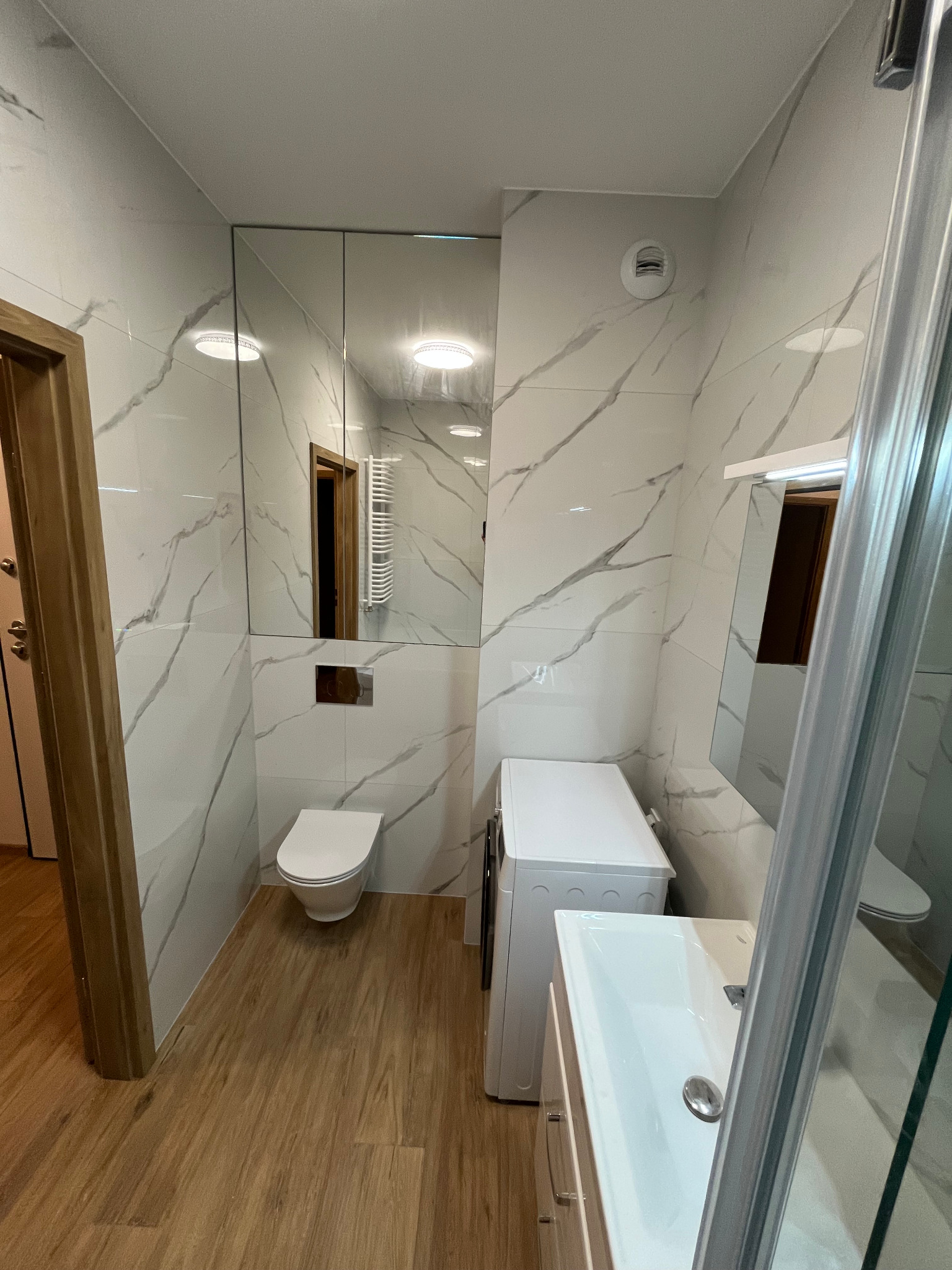 Nowoczesna łazienka z marmurowymi płytkami, drewnopodobną podłogą, toaletą, pralką i umywalką z szafką, widoczna przez częściowo otwarte drzwi z drewnianą framugą.