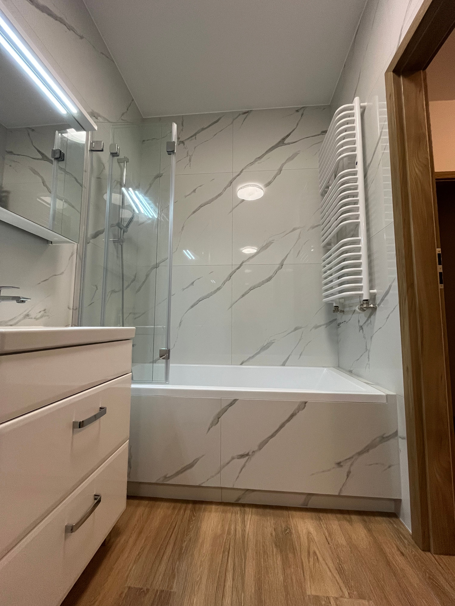 Nowoczesna łazienka z wanną obudowaną płytkami imitującymi marmur, szklanym parawanem nawannowym, białą szafką umywalkową z chromowanymi uchwytami i grzejnikiem drabinkowym, na podłodze panele...