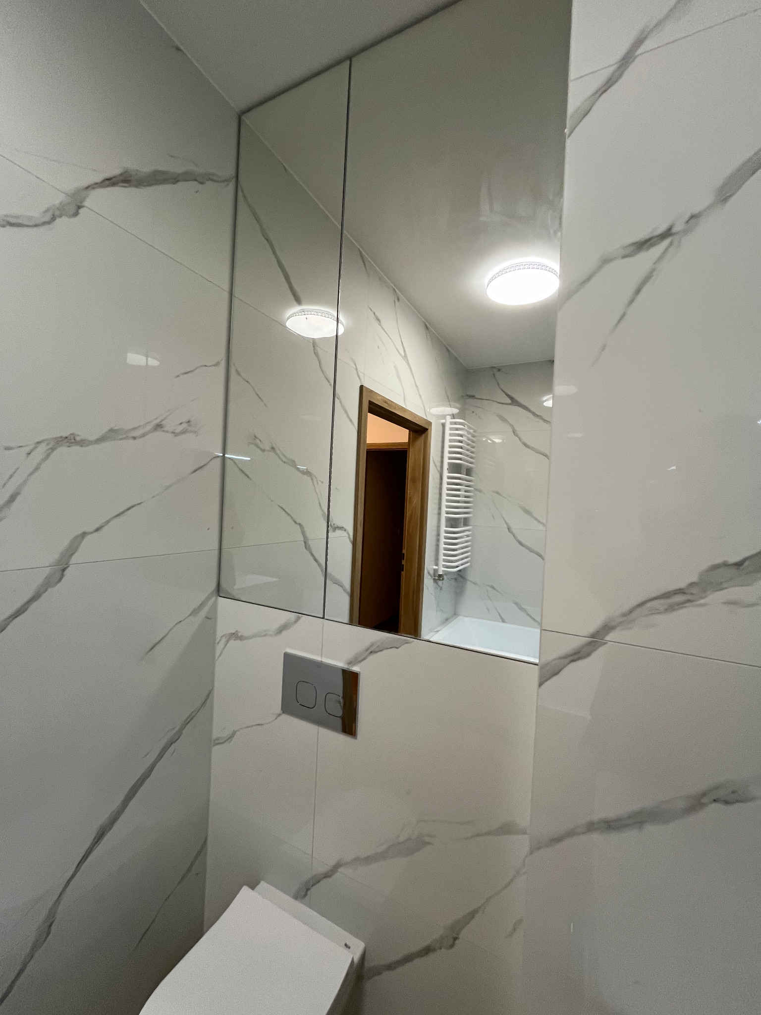 Nowoczesna łazienka z białymi płytkami imitującymi marmur, wisząca toaleta i lustro w szafce, w którym odbija się widok na wannę i grzejnik drabinkowy.