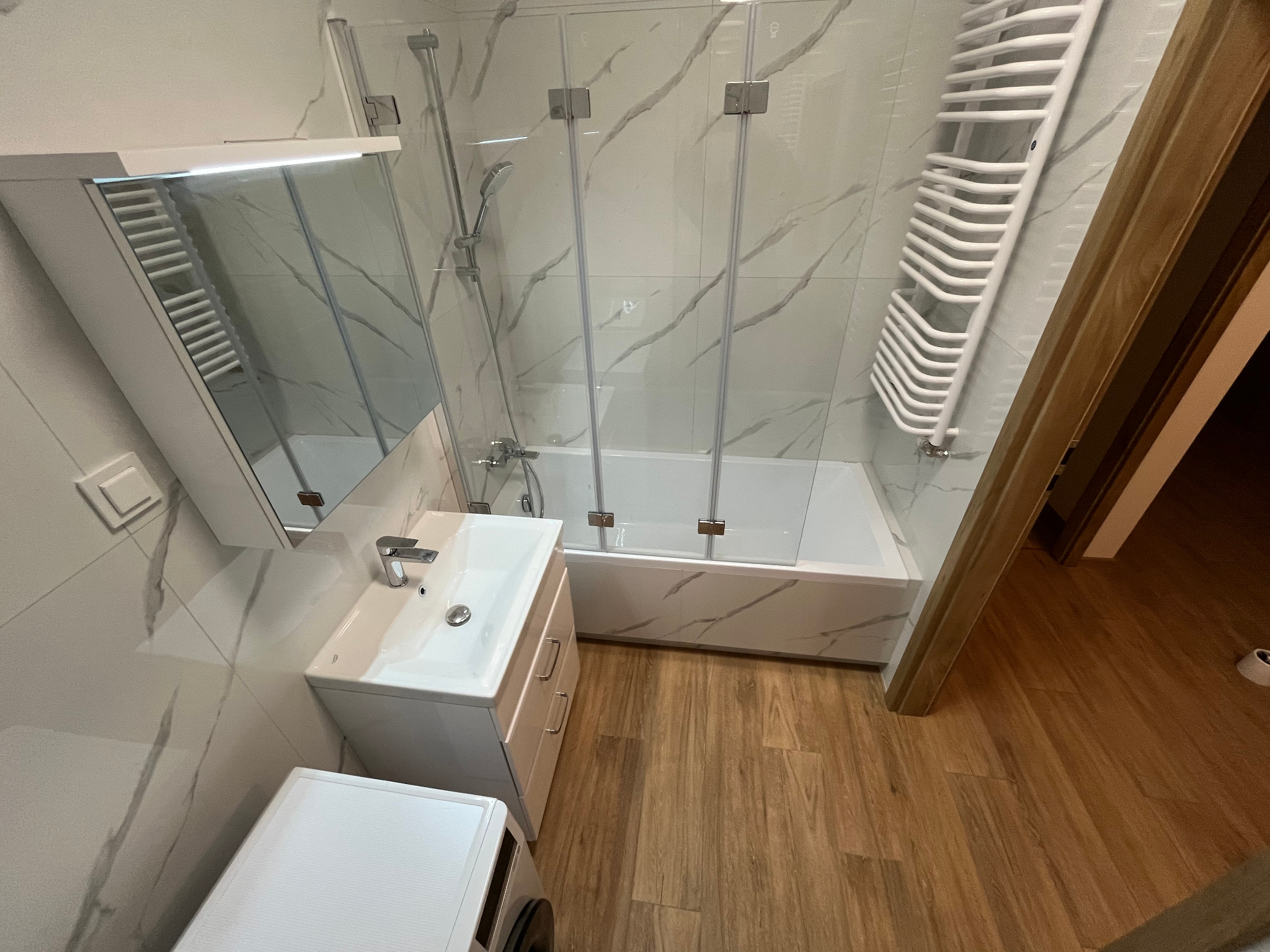 Nowoczesna łazienka z wanną za szklanym parawanem, białą umywalką z szafką, grzejnikiem drabinkowym i marmurowymi płytkami na ścianach, uzupełniona drewnopodobną podłogą.