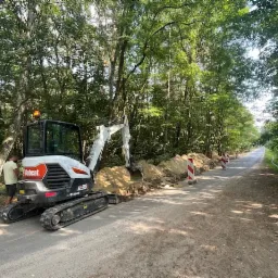 Bobcat E35 mini koparka podczas prac ziemnych przy drodze, widoczne wykopane rowy i słupki ostrzegawcze, w tle las.