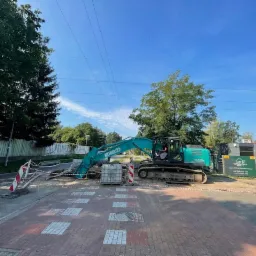 Niebieska koparka Kobelco na tle wykopu zabezpieczonego barierkami, kostka brukowa i budynek w tle, słoneczny dzień.