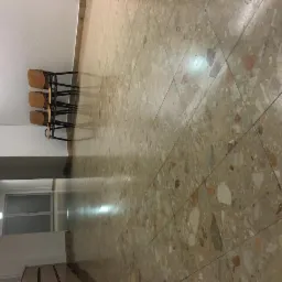 Hall z podłogą wyłożoną terazzo w odcieniach beżu i brązu, trzy krzesła ustawione pod ścianą, widoczny fragment korytarza z przeszklonymi drzwiami.