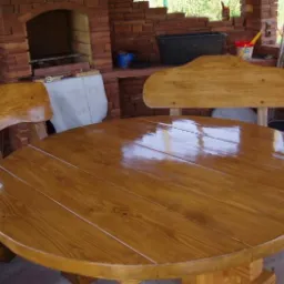 Drewniany stół i krzesła w altanie z ceglanym grillem w tle. Całość utrzymana w ciepłej, naturalnej kolorystyce, z widocznym połyskiem lakieru na meblach.