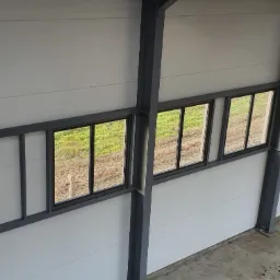 Wnętrze hali w trakcie budowy z widoczną konstrukcją stalową, oknami i płytami warstwowymi na ścianach, oświetlone światłem dziennym wpadającym przez świetlik dachowy.