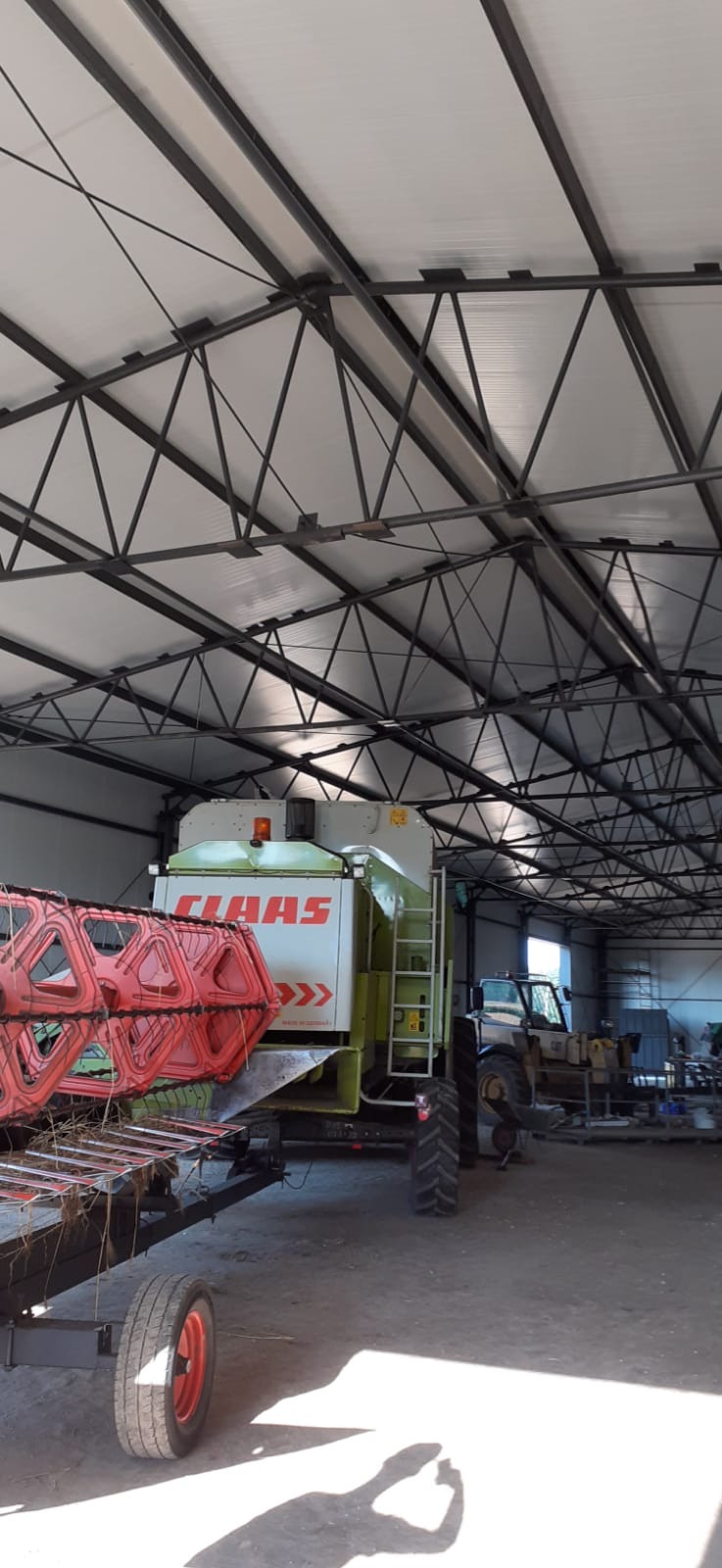 Wnętrze hali z konstrukcją stalową, w której stoi kombajn zbożowy marki Claas z czerwonym hederem, widoczny również ciągnik rolniczy.