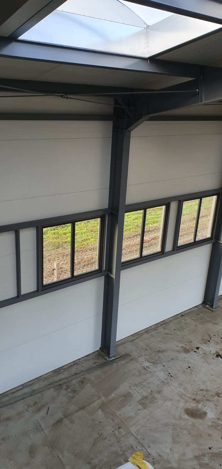 Wnętrze hali w trakcie budowy z widoczną konstrukcją stalową, oknami i płytami warstwowymi na ścianach, oświetlone światłem dziennym wpadającym przez świetlik dachowy.