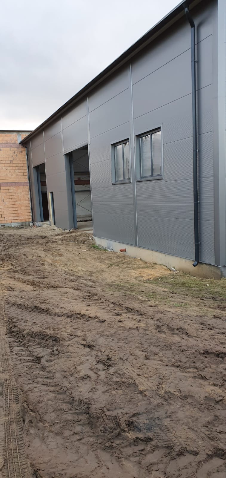 Szara hala przemysłowa z dwoma otworami bramowymi i dwoma oknami, obok budynku z czerwonej cegły, teren wokół pokryty błotem z widocznymi śladami opon.