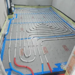 Białe rury ogrzewania podłogowego ułożone na szarej macie z niebieską taśmą, widoczne fragmenty ścian w surowym stanie.