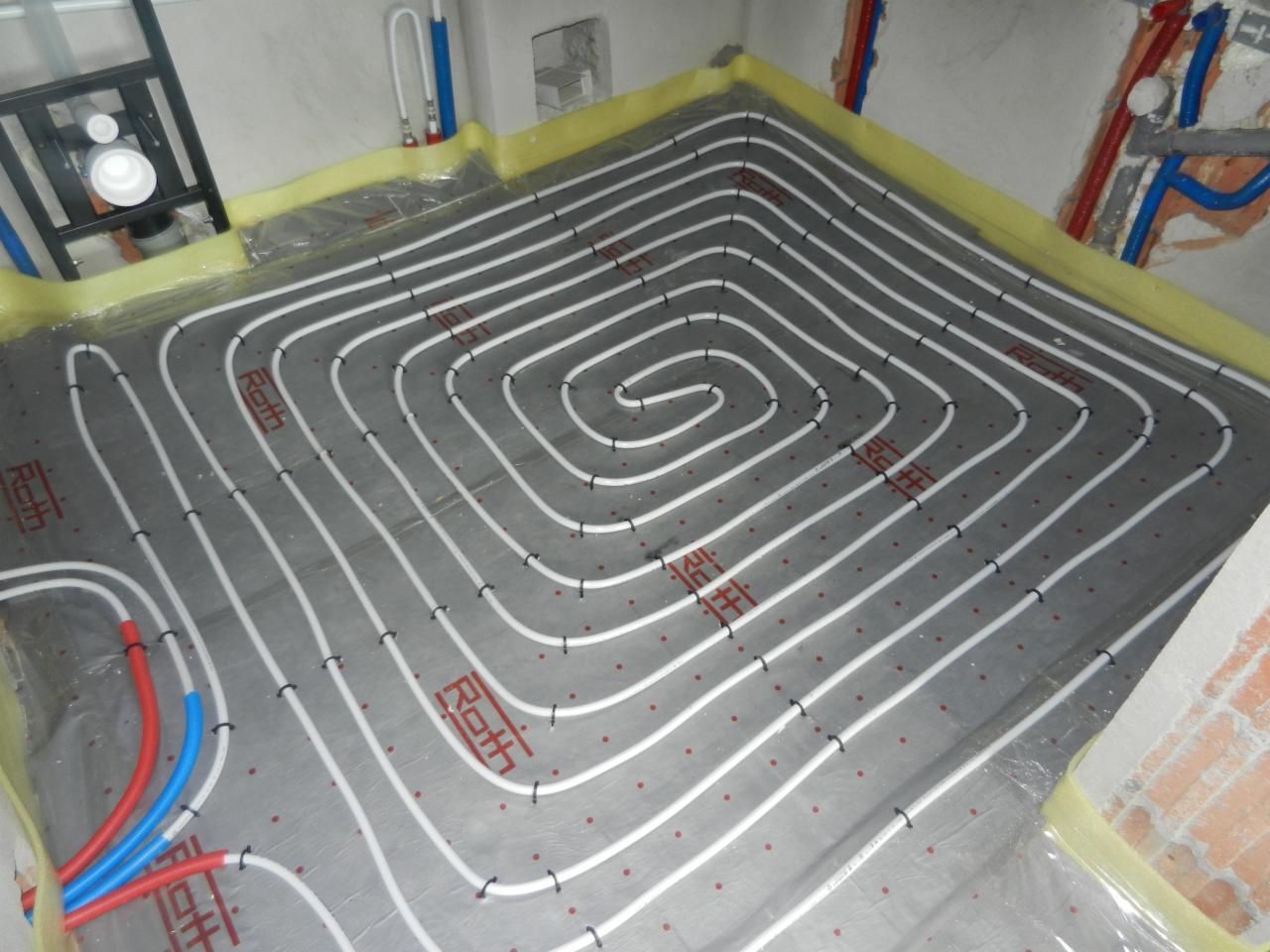 Białe rury ogrzewania podłogowego ułożone spiralnie na szarej folii z widocznymi czerwonymi punktami mocowania, żółtą taśmą brzegową i fragmentami instalacji.