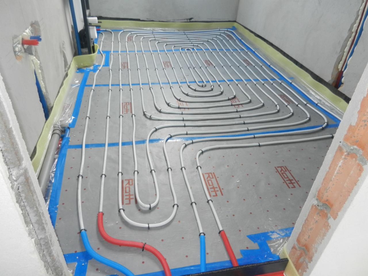 Białe rury ogrzewania podłogowego ułożone na szarej macie z niebieską taśmą, widoczne fragmenty ścian w surowym stanie.