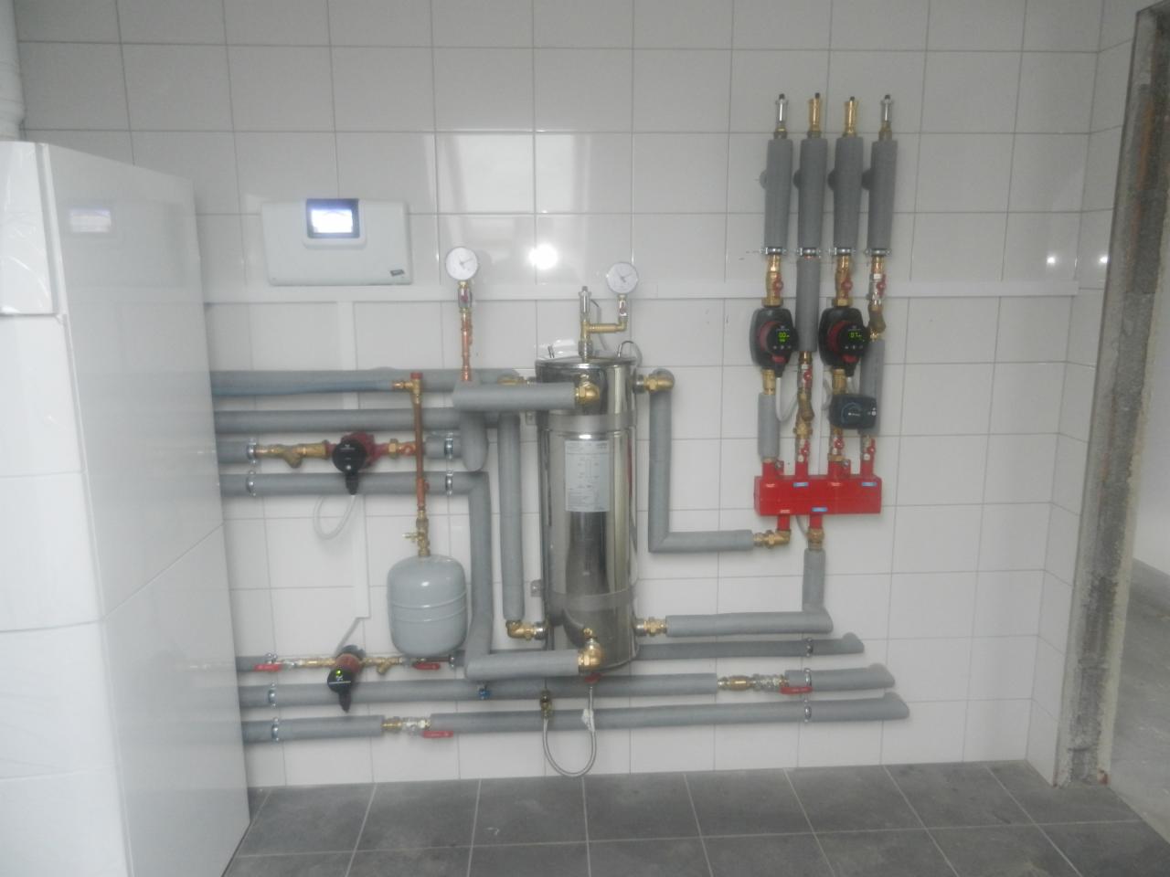 Instalacja grzewcza z pompą ciepła w kotłowni, rury w izolacji, manometry i zawory. Realizacja w Poznaniu. Białe płytki na ścianach i szara podłoga.