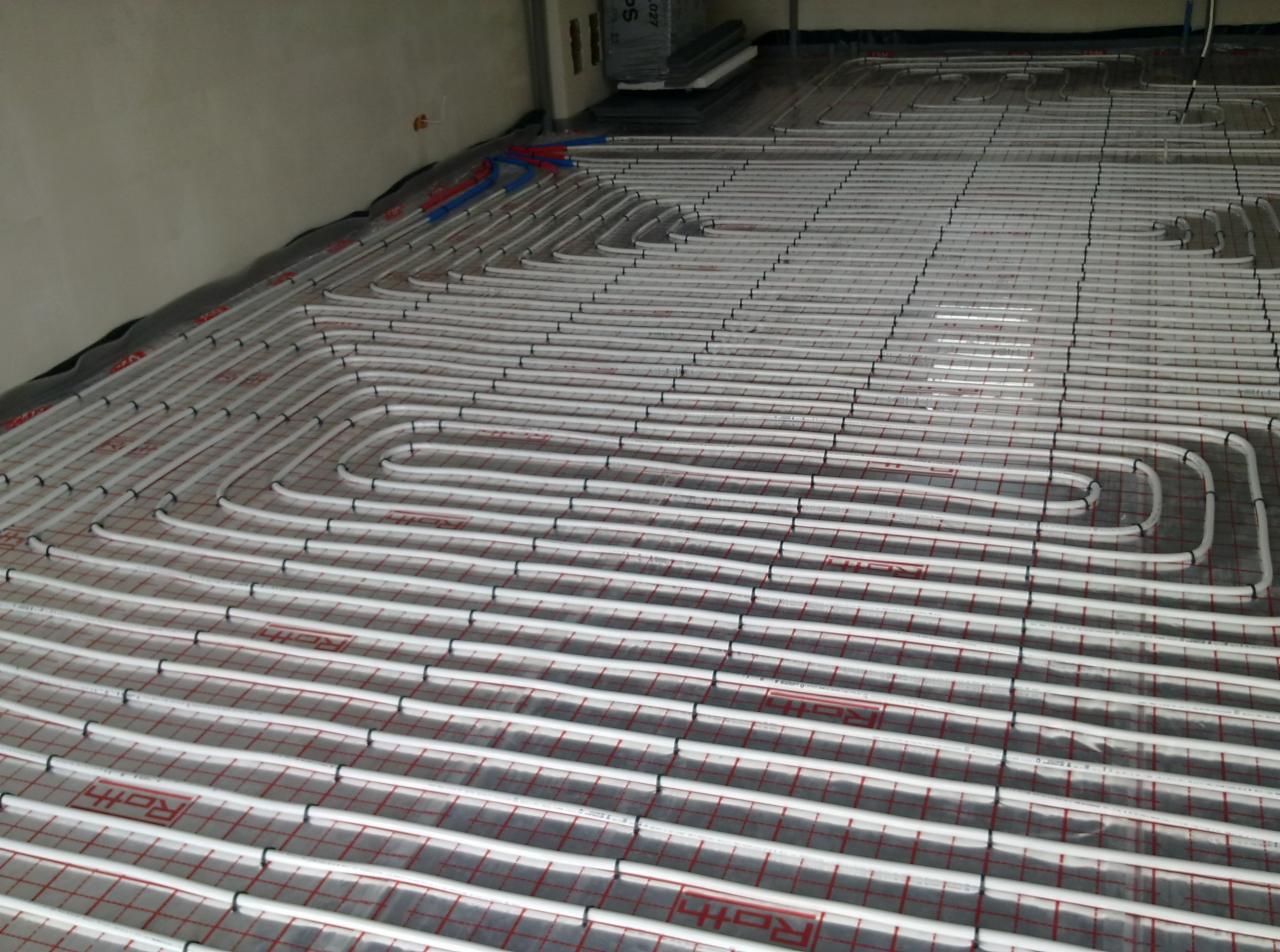 Białe rury ogrzewania podłogowego ułożone na folii z czerwoną siatką, przymocowane czarnymi uchwytami. Widok z góry na instalację przed zalaniem posadzką.