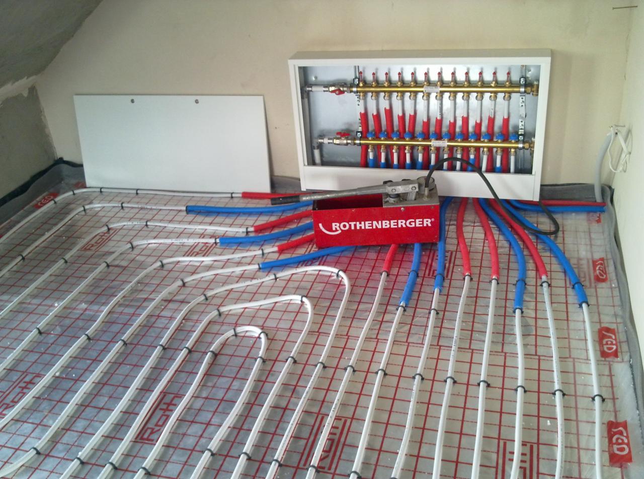 Rozdzielacz i rury ogrzewania podłogowego w kolorach czerwonym i niebieskim, ułożone na siatce. Widoczny sprzęt Rothenberger. Ściana w tle surowa.