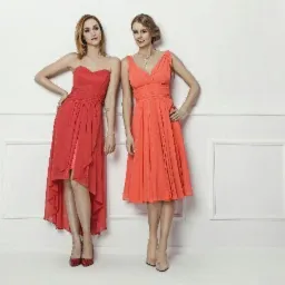 Dwie kobiety w eleganckich, zwiewnych sukienkach w odcieniach koralu i czerwieni, pozujące na tle białej ściany z dekoracyjnymi panelami.