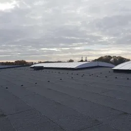 Widok z góry na płaski dach pokryty papą z widocznymi łączeniami i punktami mocowania, z dwoma świetlikami o łukowatym kształcie, w tle zabudowania i pochmurne niebo.