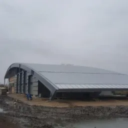 Budowa hali z łukowym dachem z blachy trapezowej w trakcie realizacji, widoczne stalowe elementy konstrukcyjne i pracownik w niebieskim kombinezonie na tle błotnistego terenu i zabudowań.