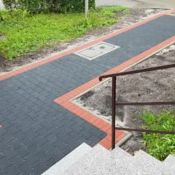 Ścieżka z szarej kostki brukowej obramowana czerwoną kostką, z wzorem w kształcie strzałki, prowadząca od schodów do trawnika, z widoczną studzienką kanalizacyjną wkomponowaną w nawierzchnię.