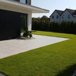 Nowoczesny taras z jasnych płyt na tle idealnie przystrzyżonego trawnika, z minimalistycznym domem w tle i ozdobnym drzewkiem bonsai.