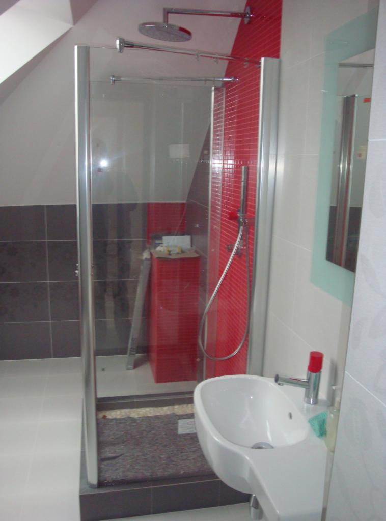 Wykończona kabina prysznicowa z czerwoną mozaiką w łazience na poddaszu. Widoczna biała umywalka i lustro z delikatną, błękitną ramą. Ukończony etap prac.