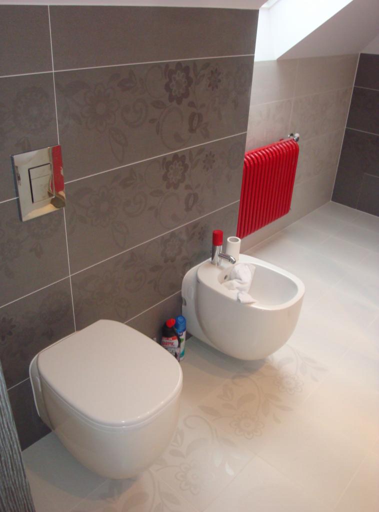 Wykończona łazienka z szarymi płytkami w kwiaty, białym bidetem i toaletą podwieszaną. Czerwony grzejnik drabinkowy i biała podłoga z delikatnym wzorem.