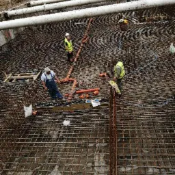 Siatka zbrojeniowa fundamentu z widocznymi rurami kanalizacyjnymi i pracownikami w odblaskowych kamizelkach, przygotowana do zalania betonem.