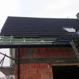 Dekarz na drabinie montuje czarną dachówkę na nowym dachu z oknem dachowym, widoczne łatowanie i fragment niezadaszonej ściany z czerwonej cegły.